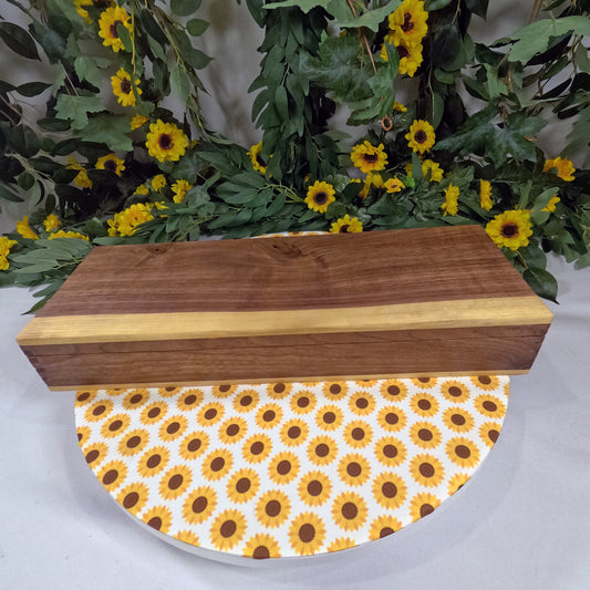 Wooden box with sunflower pattern on a white surface, surrounded by greenery and yellow flowers.